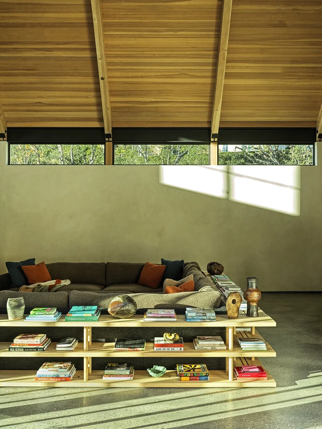 Living room of Canyon House with vaulted wood ceiling and minimal furnishings, showing a sectional sofa and a low wooden bookshelf filled with books.