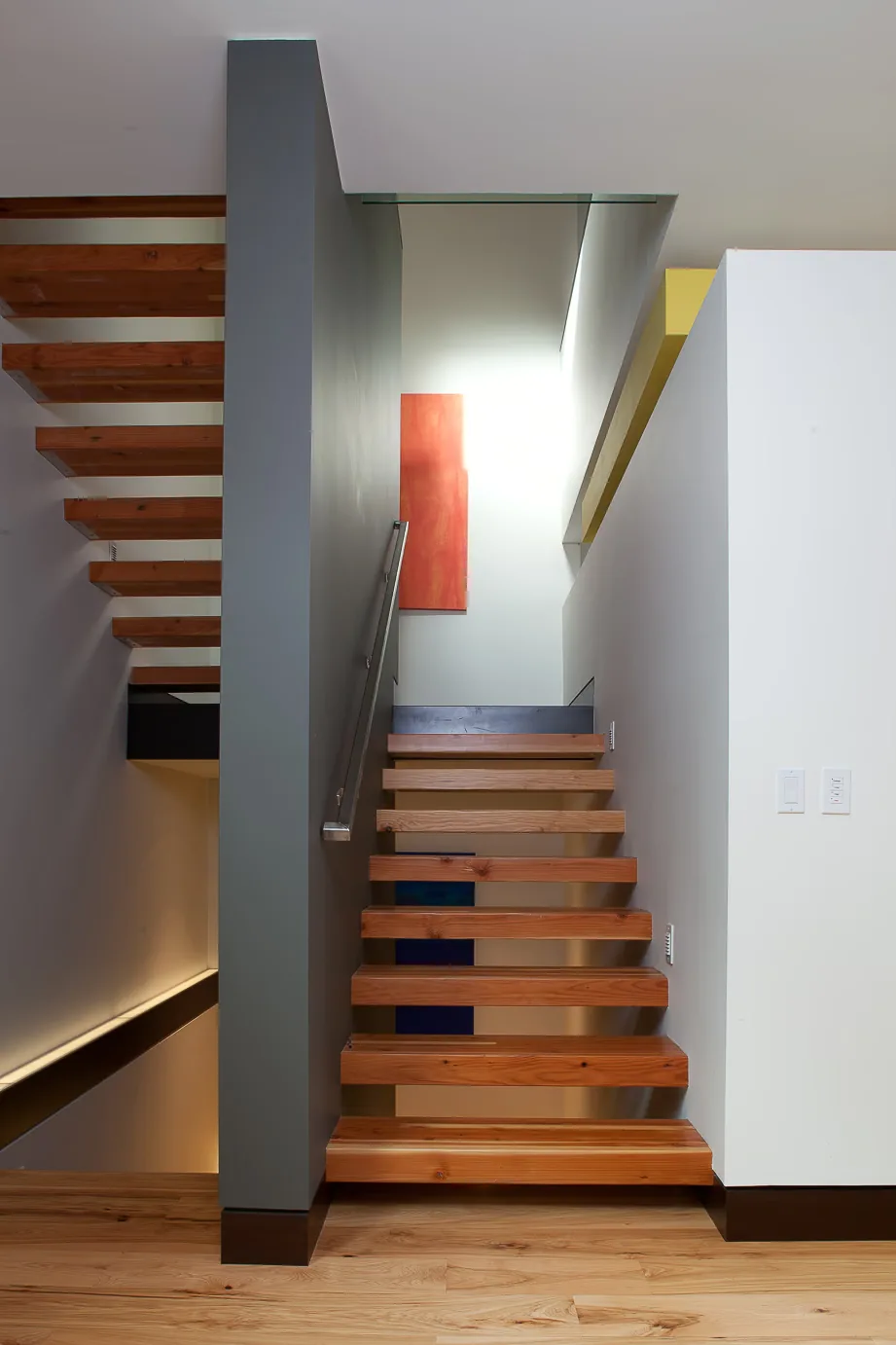 Floating wooden stairs with a metal handrail lead upward alongside a gray wall in a contemporary interior.