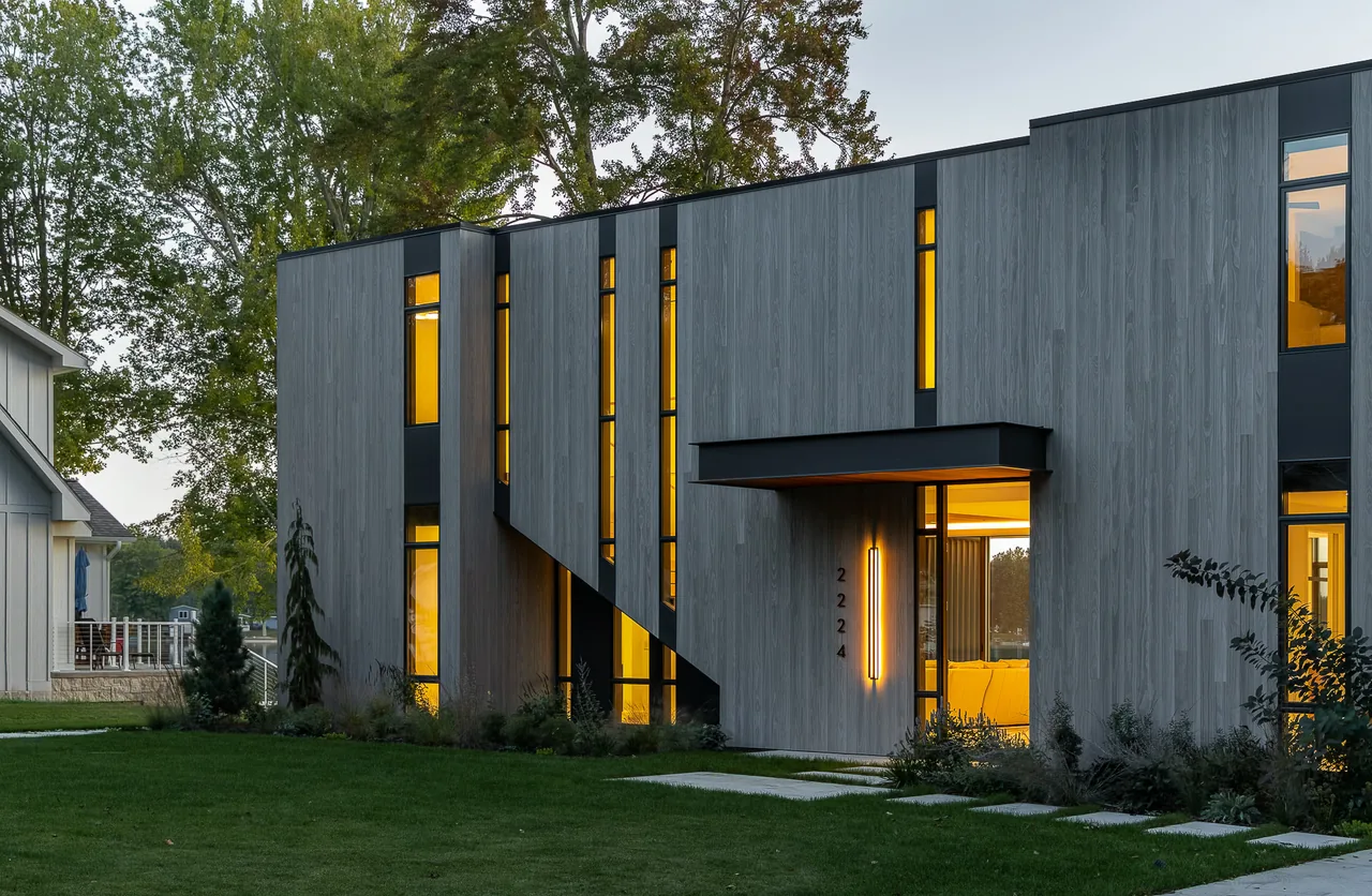 Hutchins Lakes Residence features vertical wood siding and tall narrow windows with black frames on the exterior front at dusk.