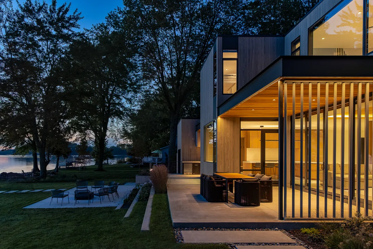 The Hutchins Lakes Residence features vertical wood siding and large glass windows overlooking a lakeside patio, lit during evening hours.