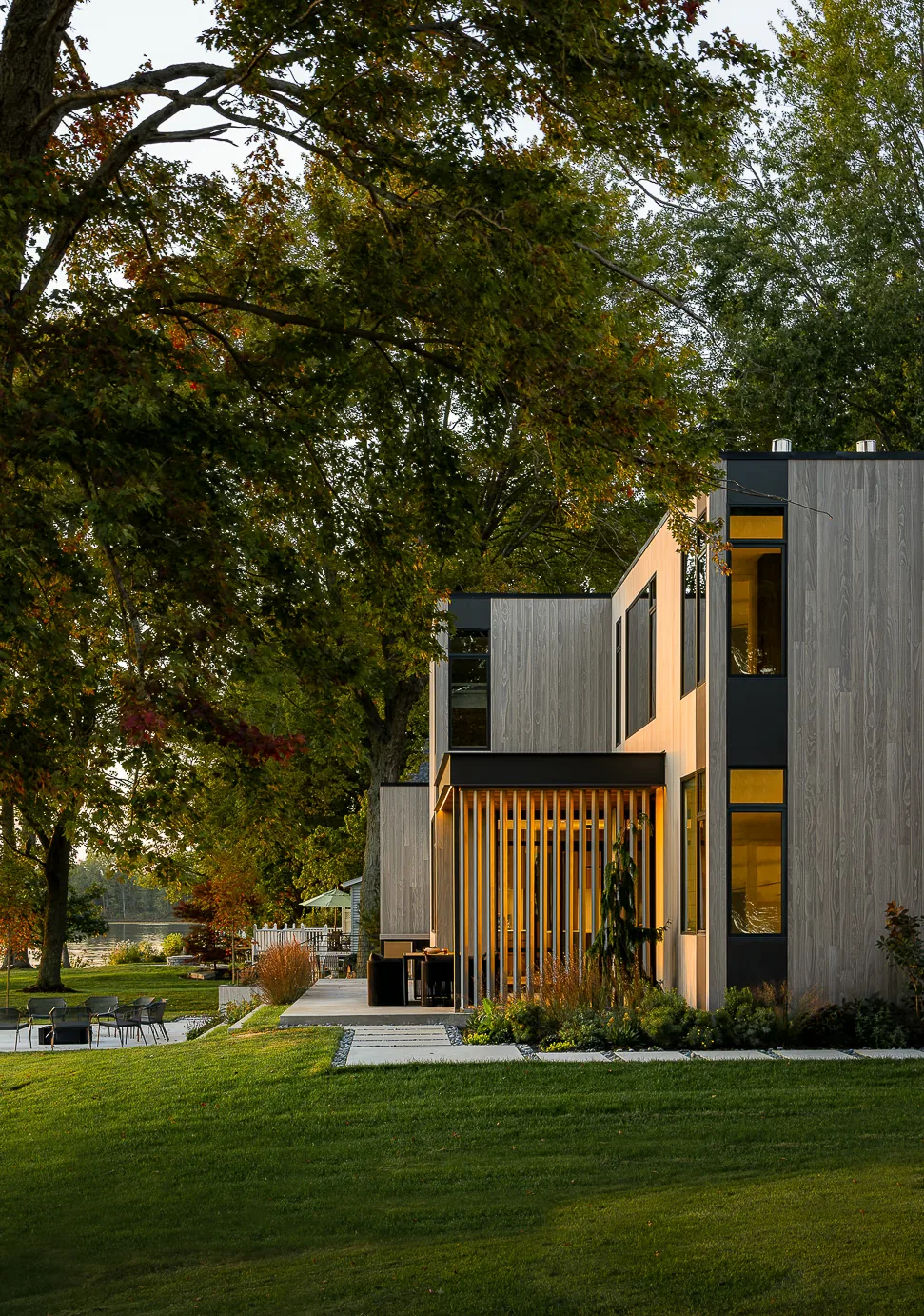 Hutchins Lakes Residence features vertical wood siding and vertical window strips with evening lighting near the lake.