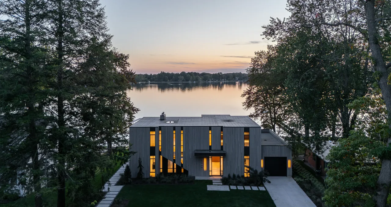 The Hutchins Lakes Residence by Lucid Architecture features a modern, rectangular design with vertical windows and wood siding by a tranquil lake.