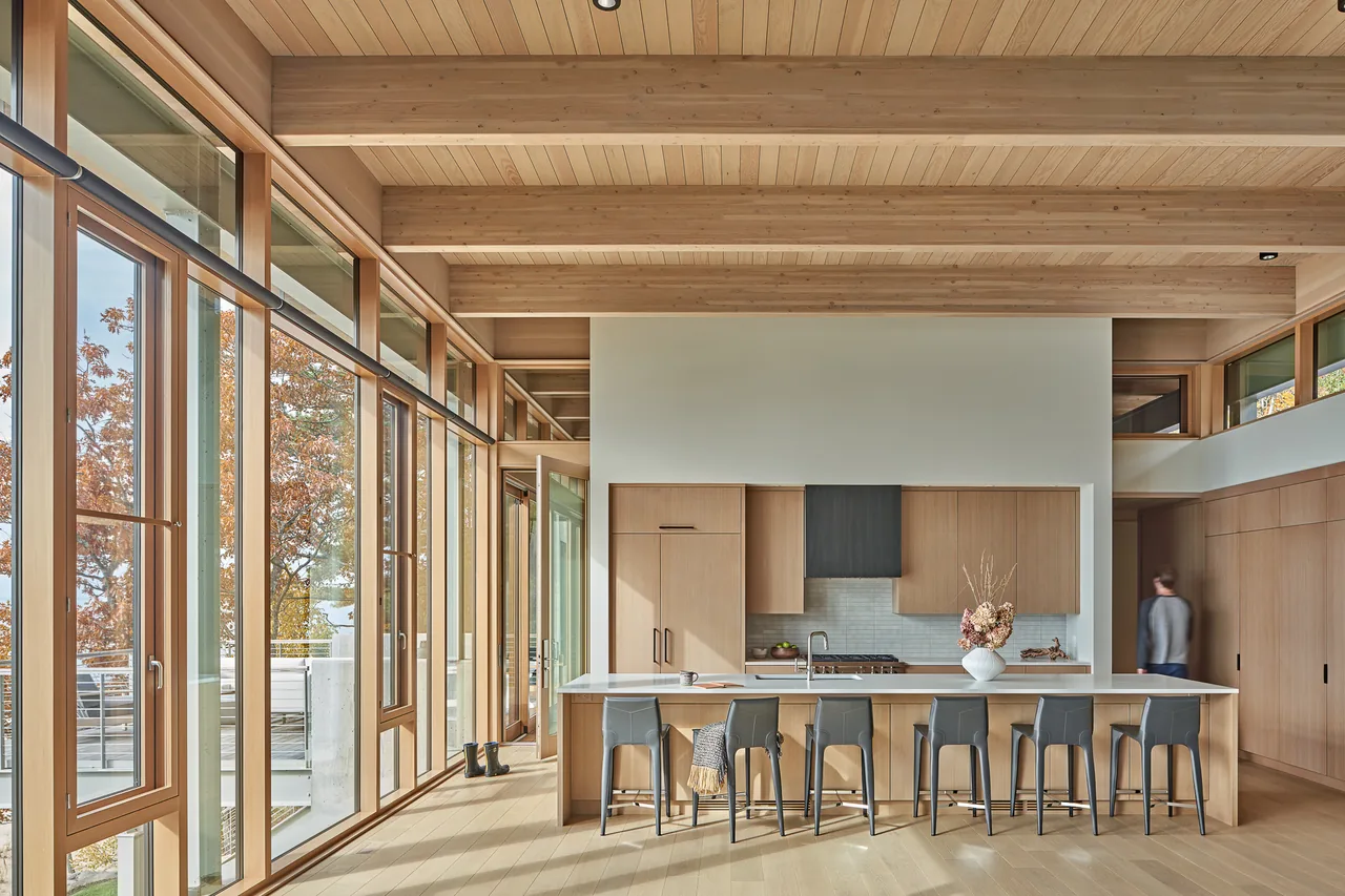 The Lower Shore Residence kitchen features wood ceiling beams, floor-to-ceiling glazing, and a kitchen island with gray bar stools.