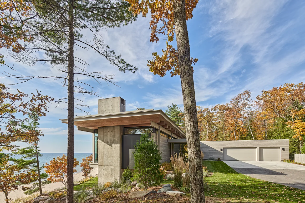 The Lower Shore Residence by Lucid Architecture features a flat roof and concrete walls with wood detailing, located near the water with autumn trees surrounding it.