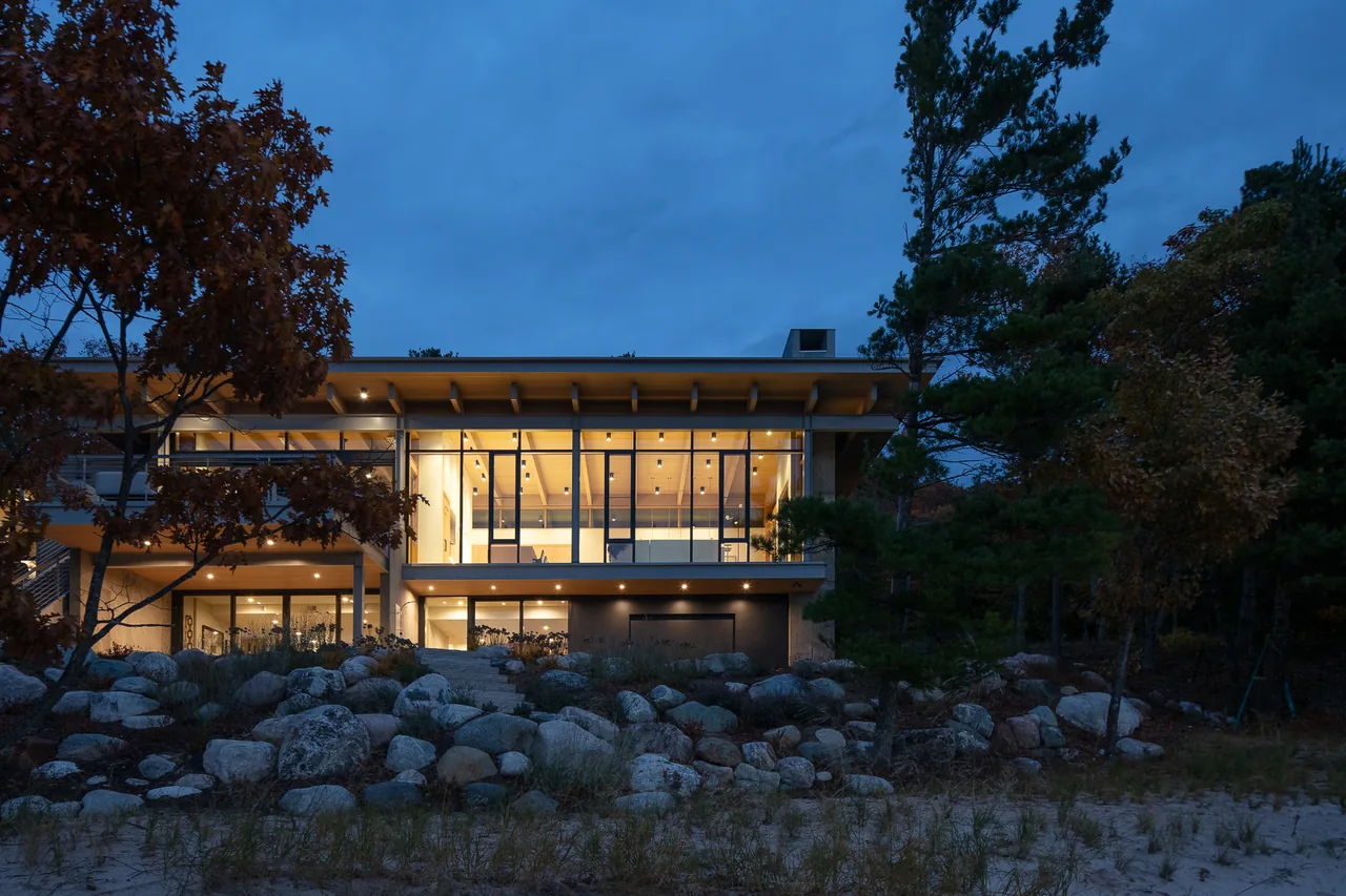The Lower Shore Residence features floor-to-ceiling glazing with exposed wood ceiling beams, blending modern design with natural rock landscaping.