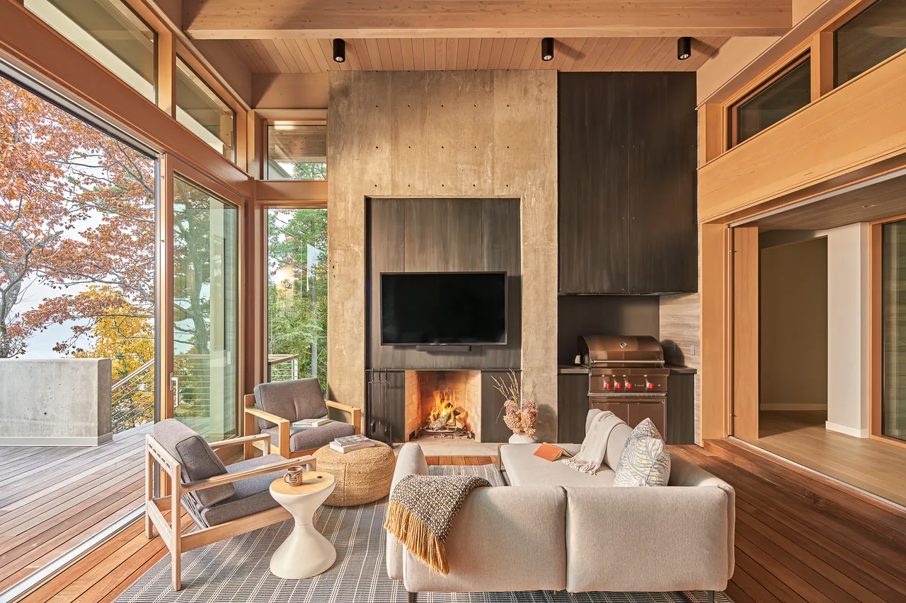 Lower Shore Residence living space features a concrete fireplace, wood ceiling, and large windows overlooking autumn foliage.