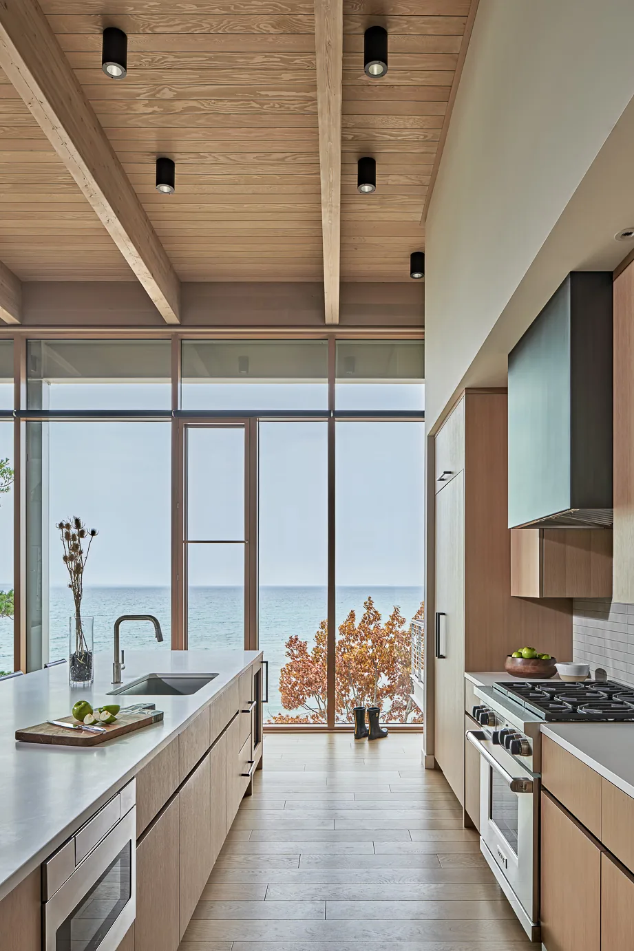 Kitchen of the Lower Shore Residence featuring wood cabinets, a countertop island, and expansive ocean views through floor-to-ceiling glazing.