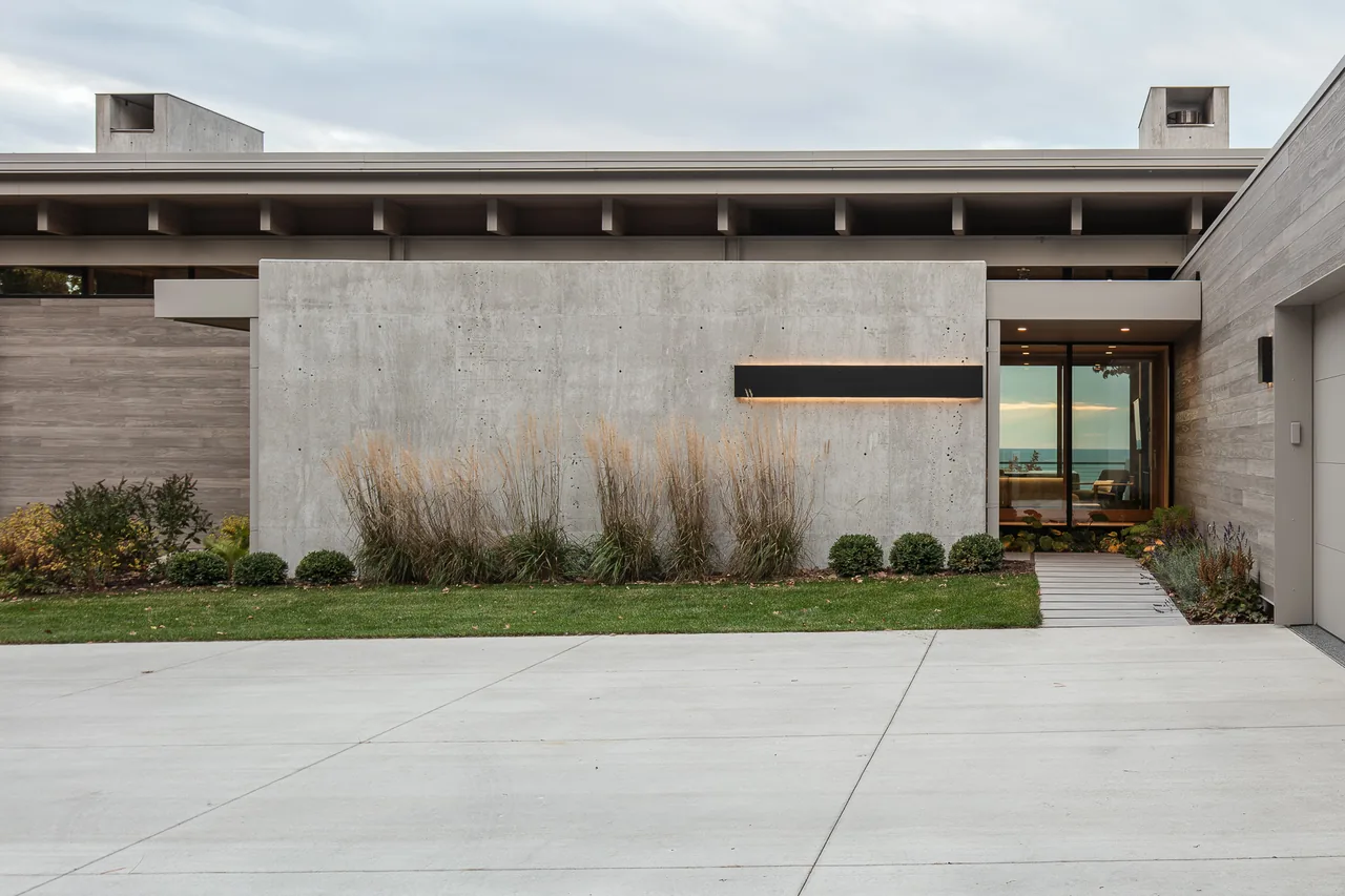 Modern residence exterior features a concrete wall, wood siding, and a glass entry door with a landscaped front.