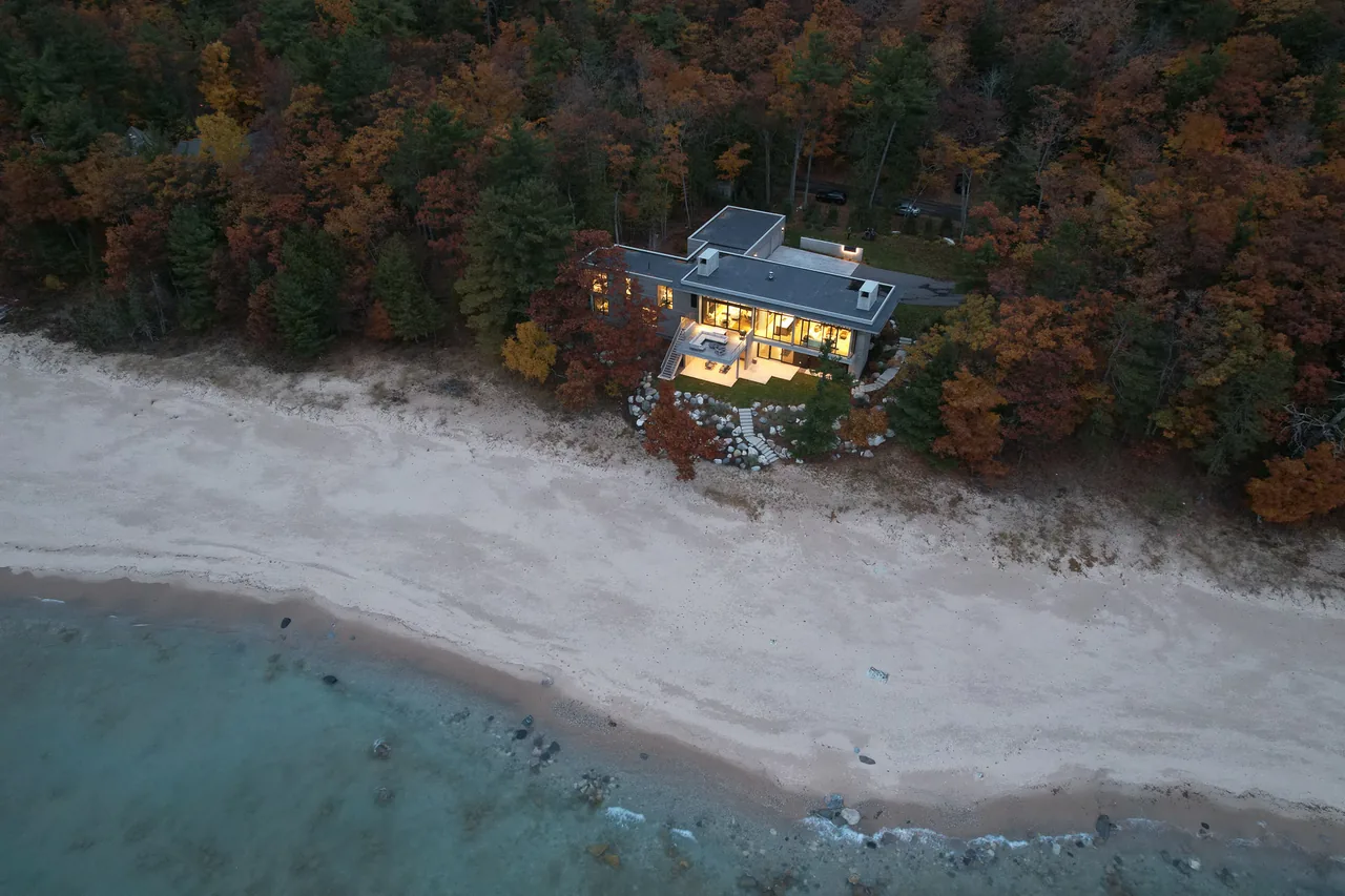 Lower Shore Residence by Lucid Architecture sits on a waterfront lot with a flat roof and large illuminated windows in autumn.