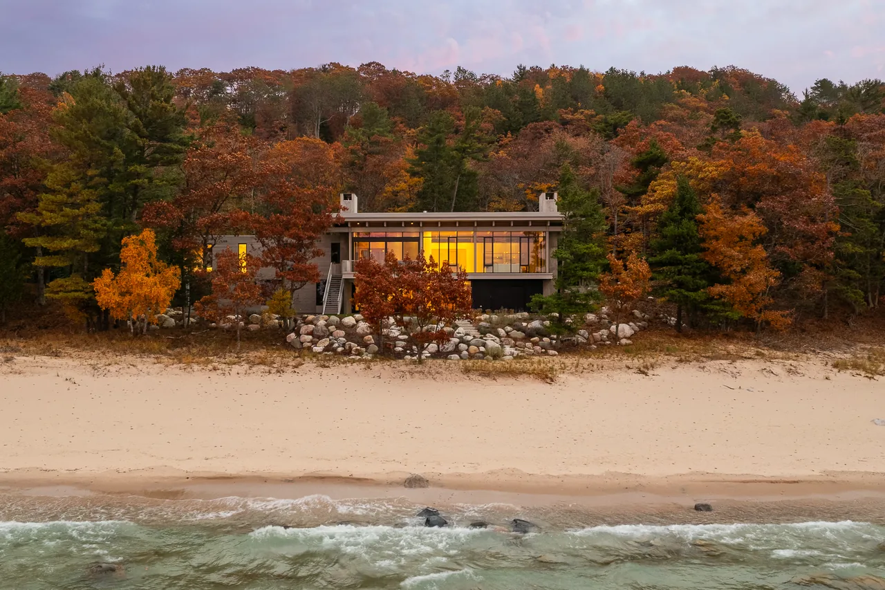 Lower Shore Residence by Lucid Architecture features floor-to-ceiling glazing and is nestled beside a sandy beach and forest with fall colors.