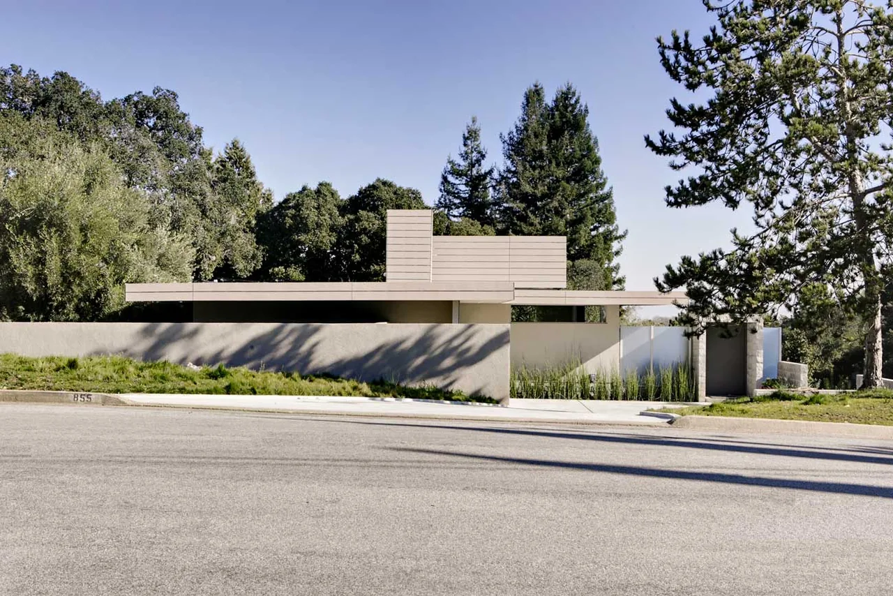 The Olson Residence features a modern design with a flat roof and horizontal wood siding, surrounded by mature trees and a low perimeter wall.