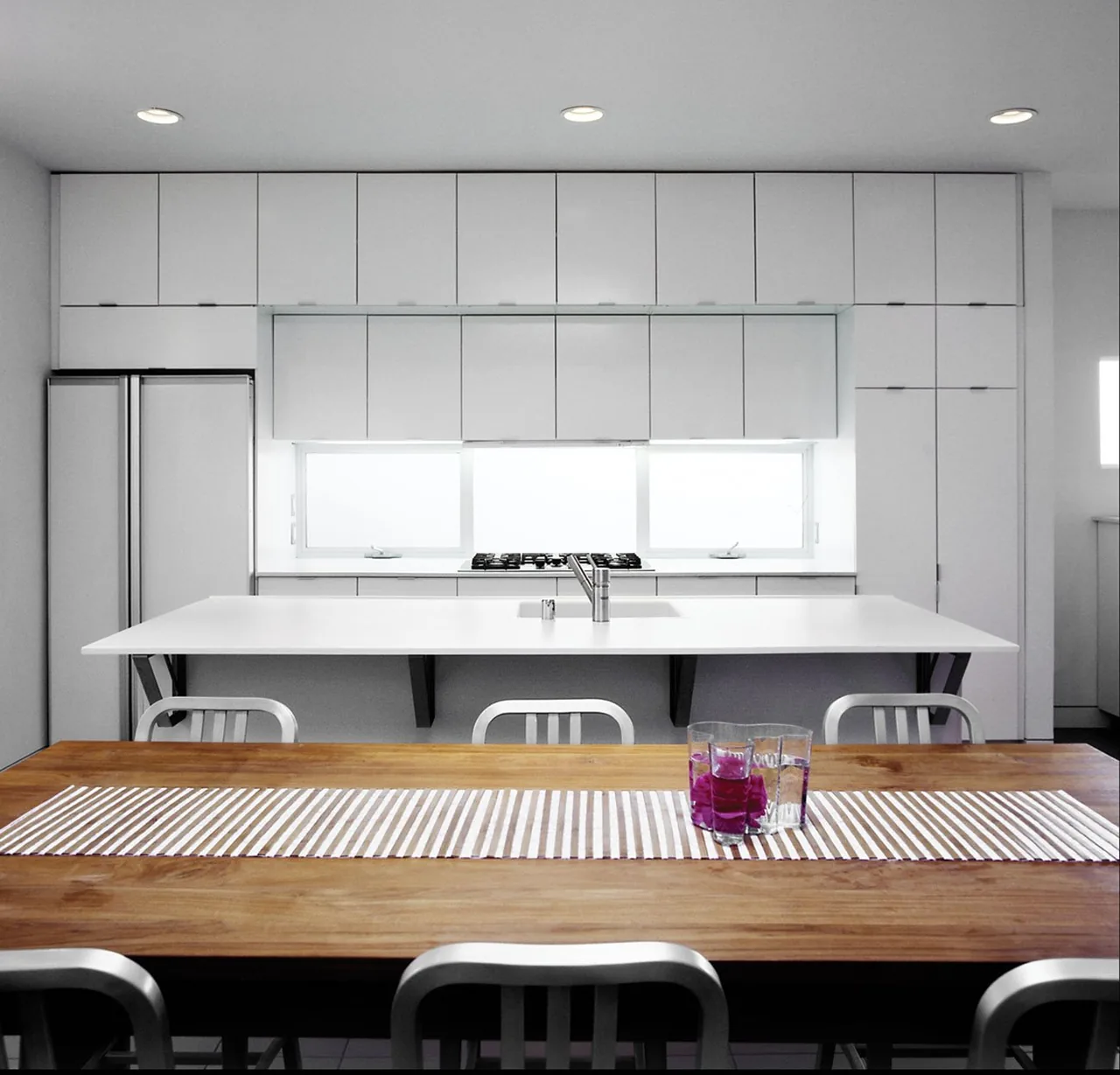 The kitchen at Cabral Residence features white cabinets, a gas cooktop below a large window, and a wood dining table with metal chairs.