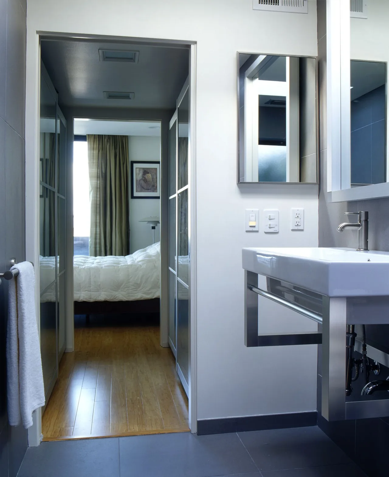 Bathroom with a stainless steel sink and mirror, showing a bedroom beyond with wood flooring and curtains.
