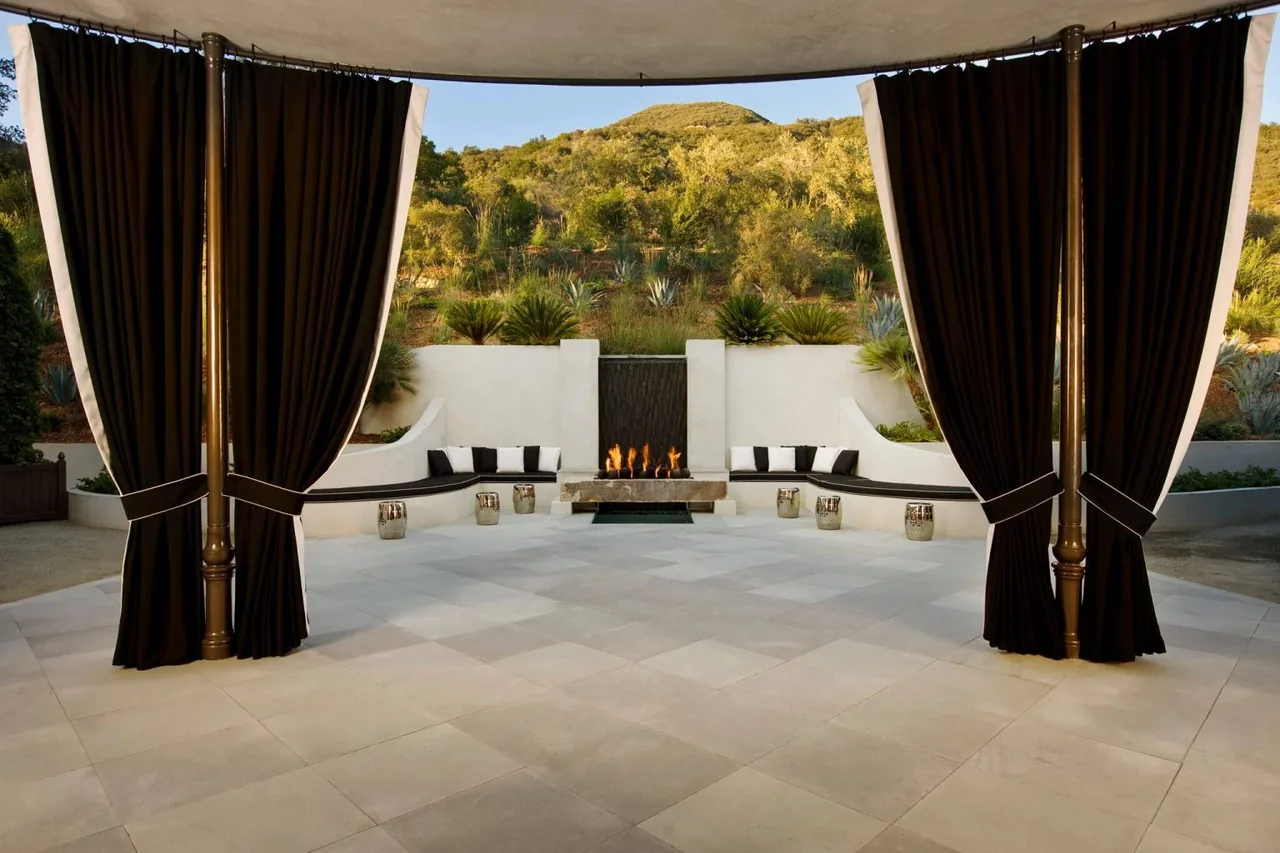 Outdoor covered patio features dark curtains, built-in curved seating with cushions, and a modern linear fireplace.