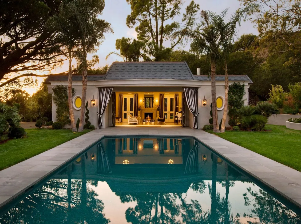 The Hollywood Regency style estate features a pool and open patio leading to an elegant interior with a fireplace.