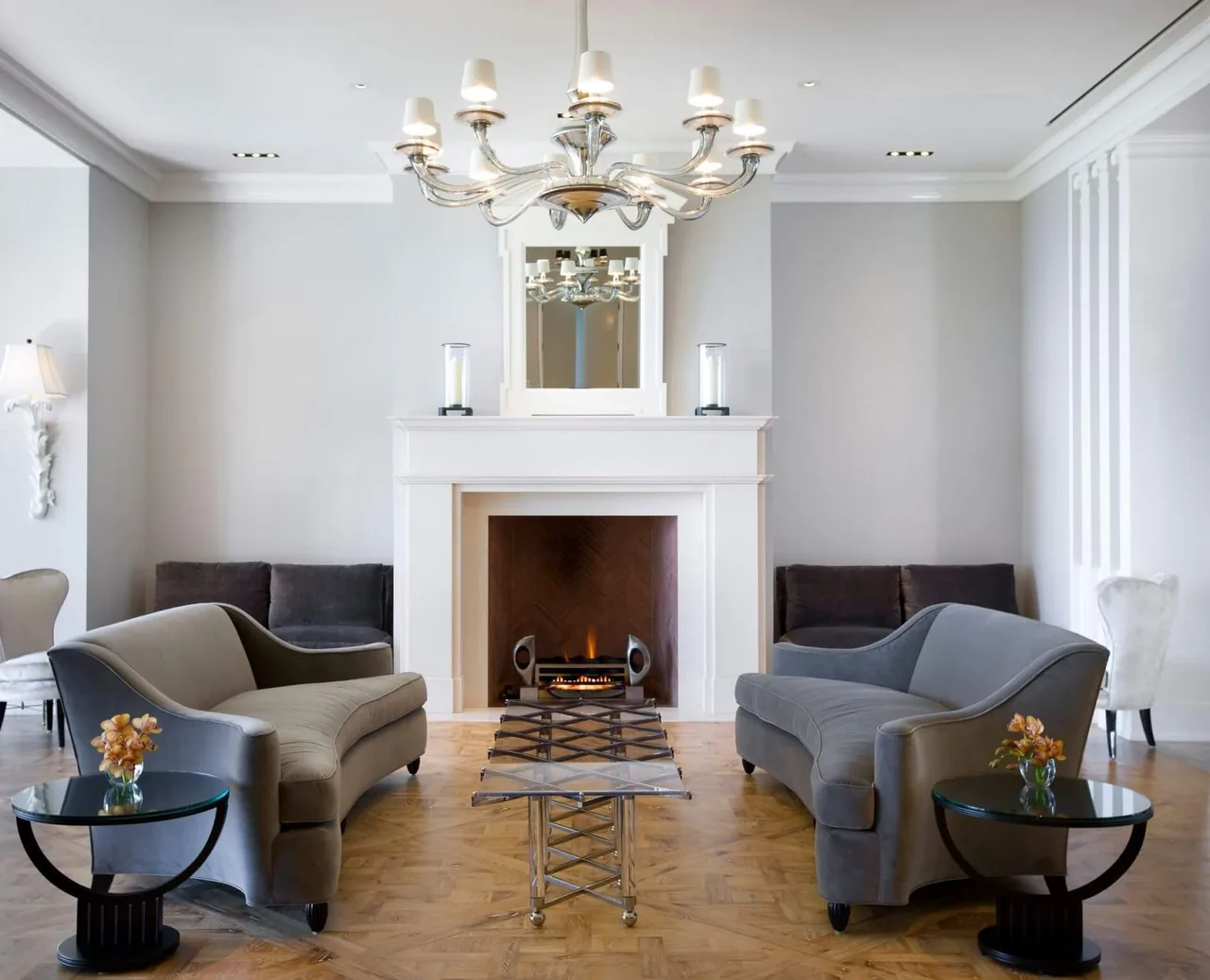 Living room features paired gray sofas, a white fireplace with a mirror above, and a chandelier, set on parquet flooring.