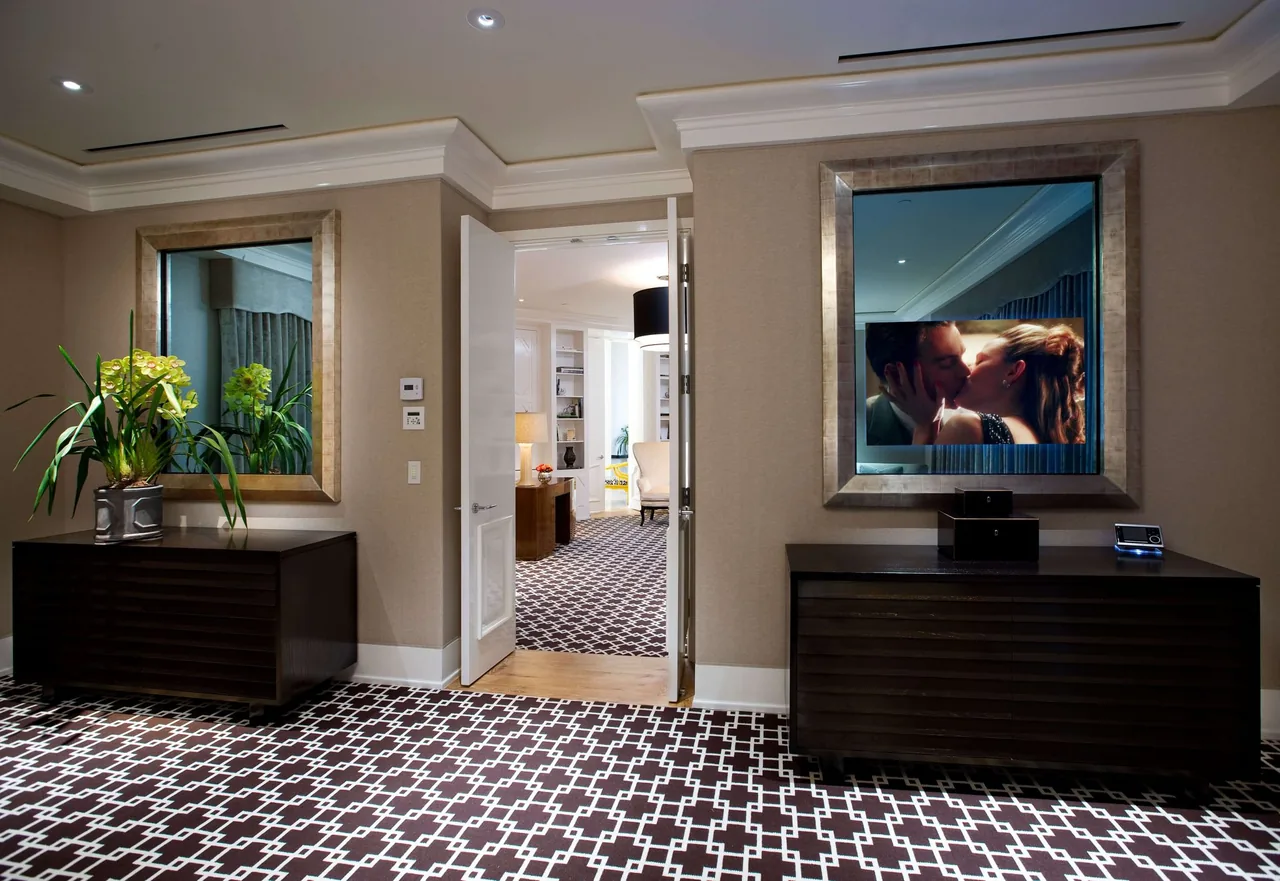 Hallway with patterned carpet, two dark wood consoles, large mirrors, and beige walls near an open doorway.