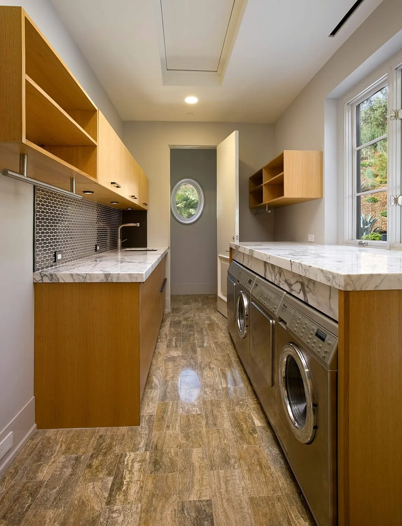 Laundry room features light wood cabinets, marble countertops, stainless steel appliances, and an oval window providing natural light.