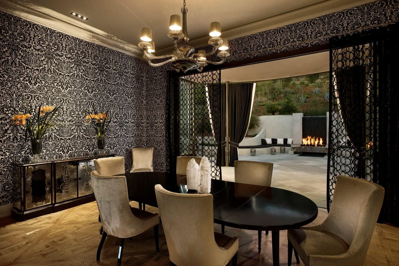 Dining room with ornate patterned wall covering, oval black table, beige upholstered chairs, and chandelier, opening to a patio with a fire feature.