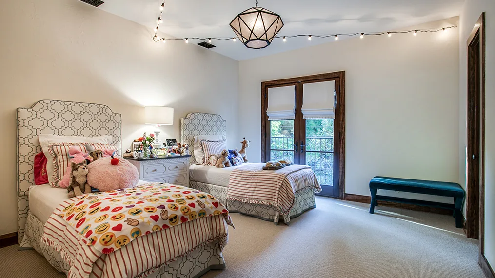 Bedroom with twin beds, each with patterned headboards and playful emoji-themed bedding, and a central nightstand with a lamp.