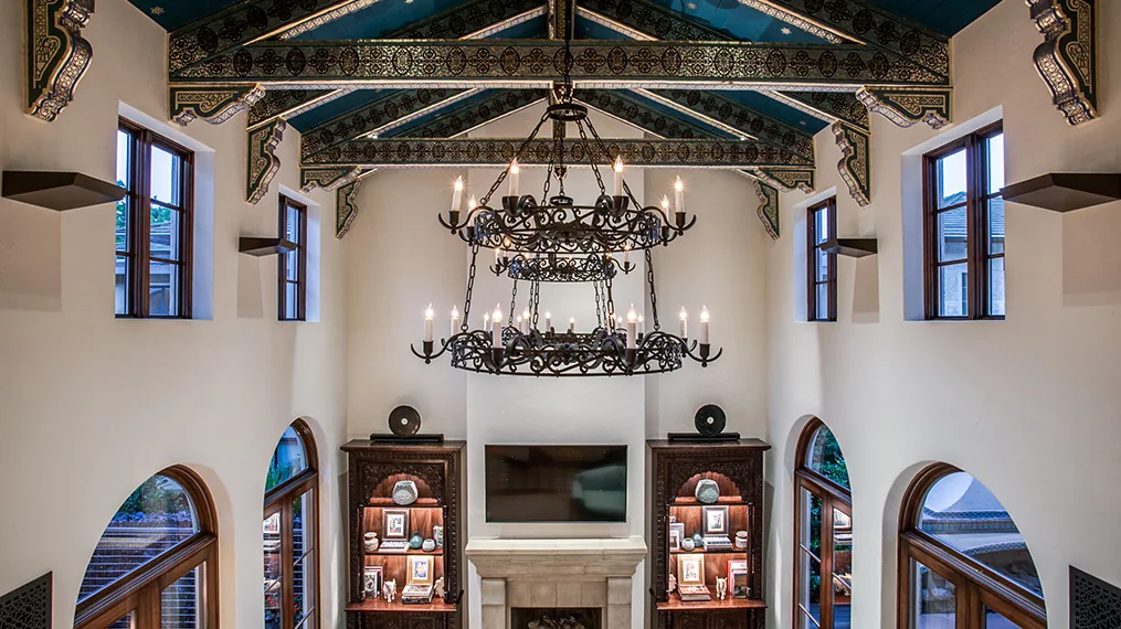 Living room with vaulted ceiling featuring ornate painted beams and two wrought iron chandeliers above a fireplace flanked by built-in shelving.