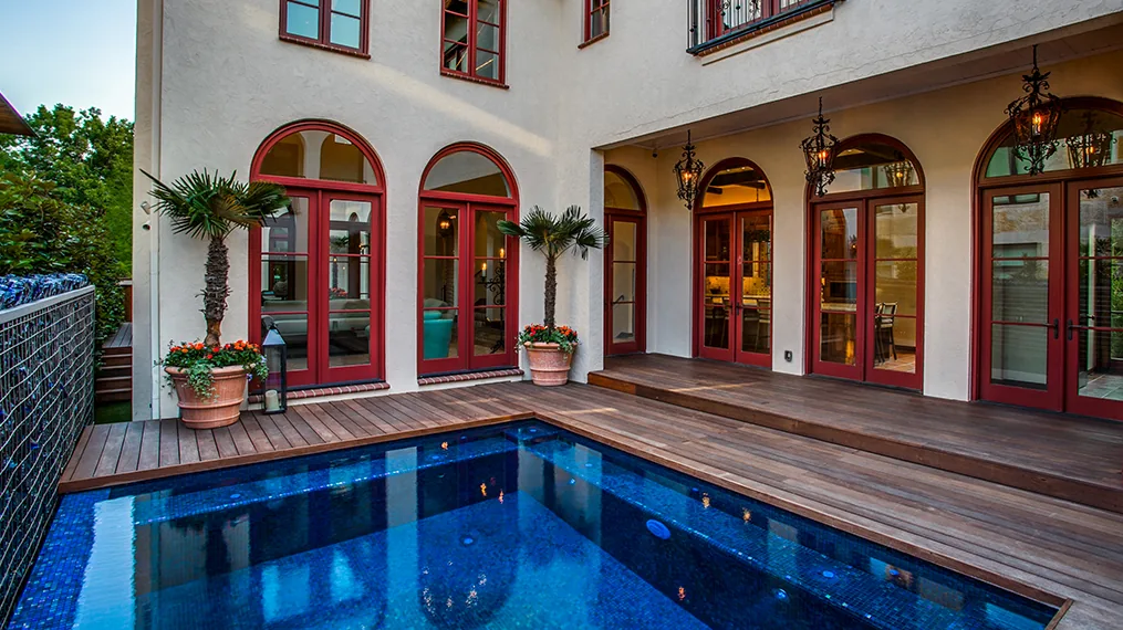 Outdoor living area with pool, wooden deck, and arched red doors and windows in Spanish Colonial Revival style home.