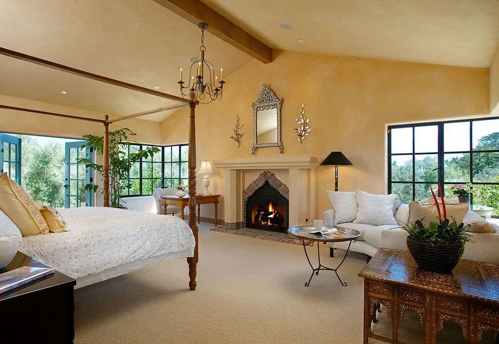 A bedroom features a four-poster bed, decorative fireplace, large windows with black frames, and a sitting area with sofa and table.
