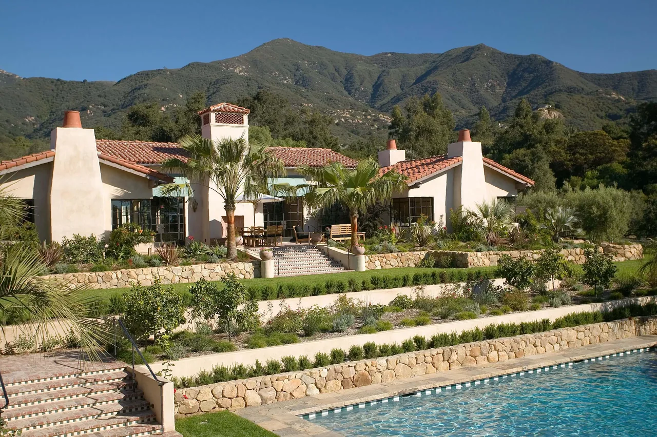 MONTECITO ANDALUSIAN ESTATE features terracotta tile roofs and stone terrace landscaping with a pool in front of mountain hills.