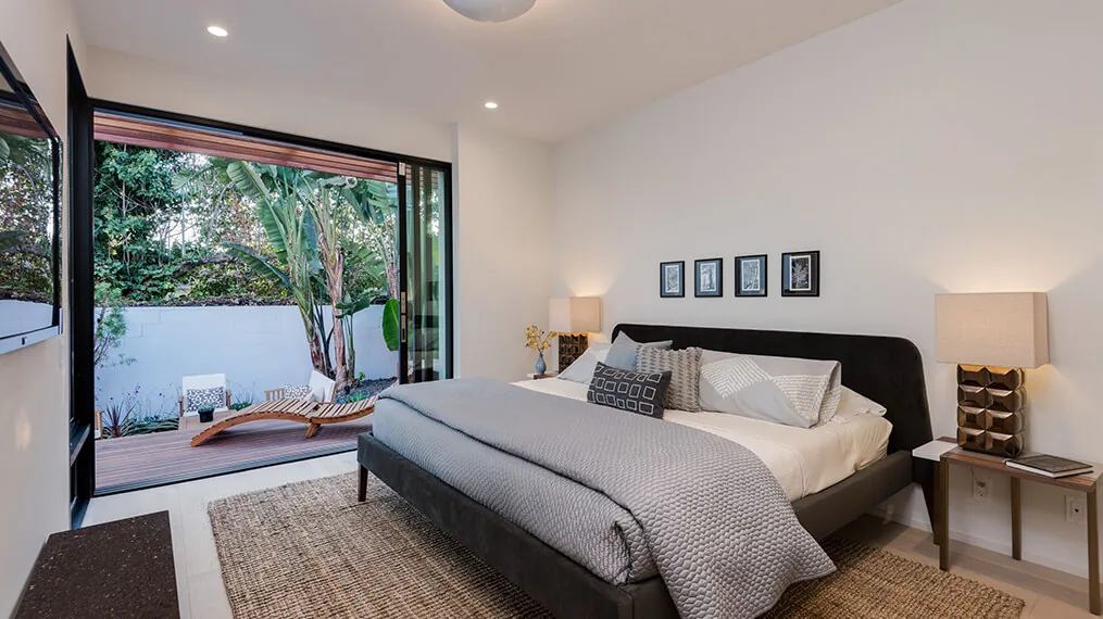 This bedroom features neutral tones, textured rug, and sliding glass doors leading to an outdoor wooden deck with lounge chairs and plants.
