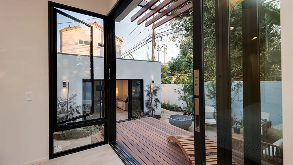 Black-framed glass doors open from interior to wooden deck with outdoor seating and planter walls.