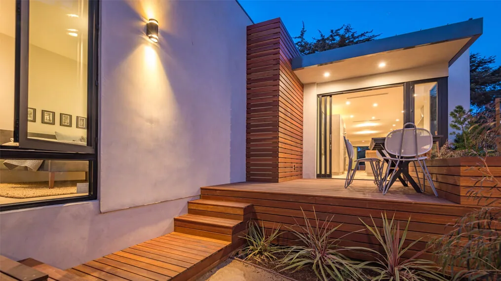 Modern bungalow exterior with wooden deck, built-in bench, and warm dusk lighting from wall sconces.