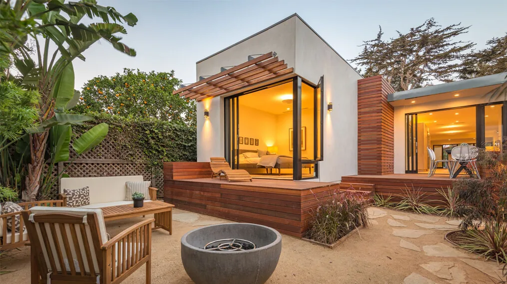 Exterior view of a modern bungalow featuring a wooden deck, outdoor seating area, and large windows revealing an illuminated bedroom interior.