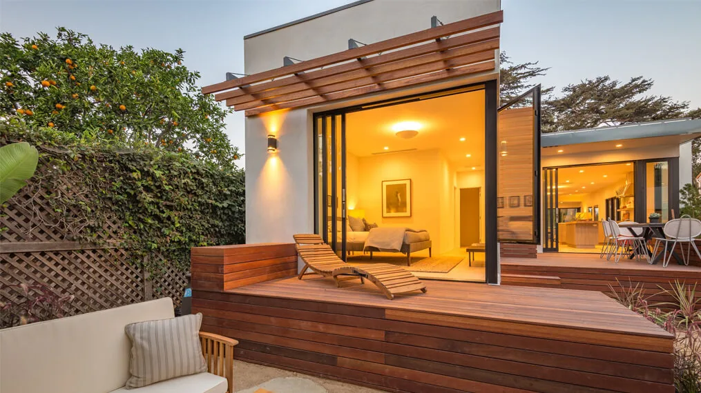 Modern bungalow exterior with wooden deck, open sliding doors, and warm interior lighting at dusk.