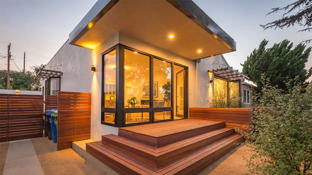 Modern bungalow exterior features a wood-slat fence, built-in bench on the patio, and black window frames illuminated by warm dusk lighting.