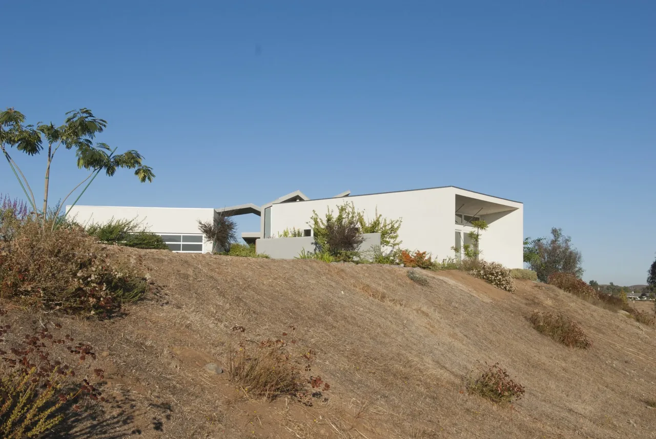 Modern white house situated on a dry hillside with angled roof and desert landscaping.