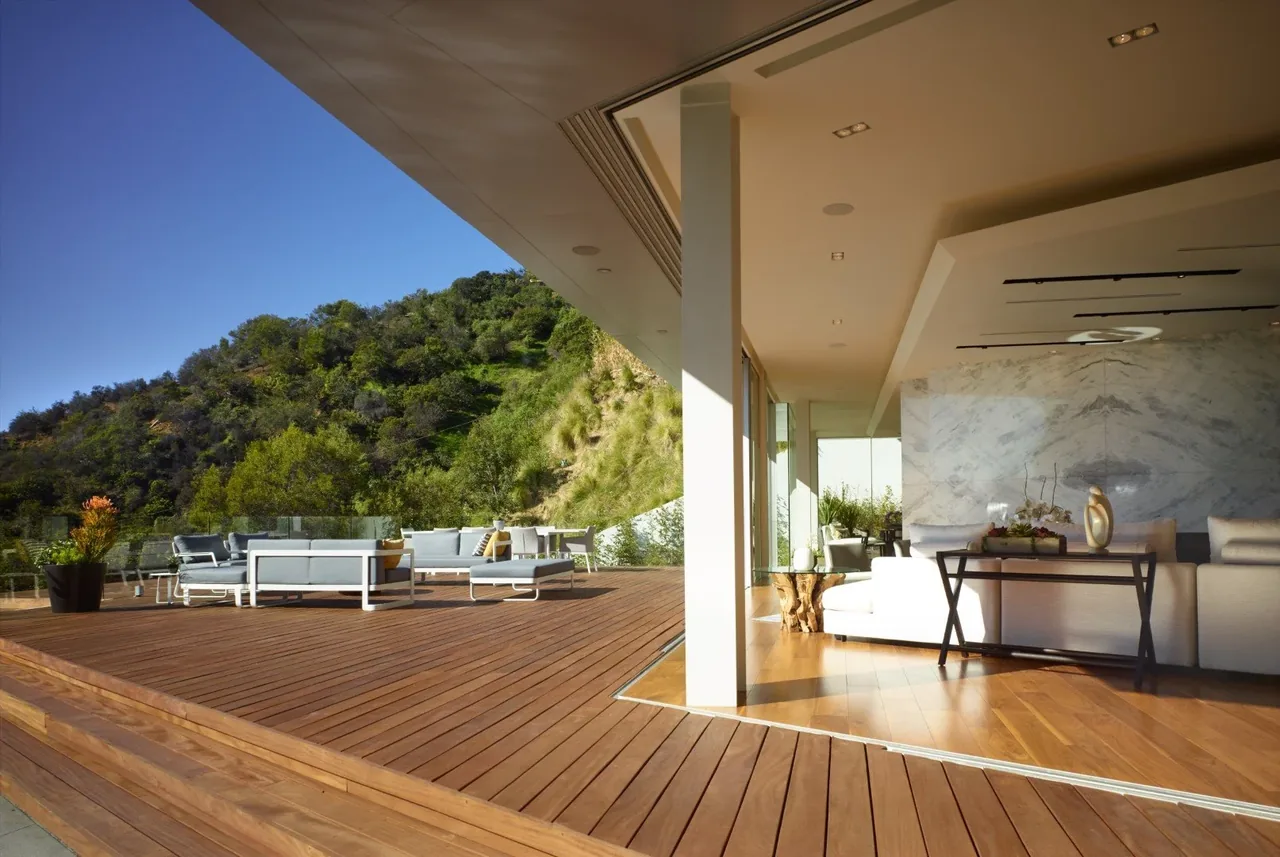 The Rising Glen project features an open living space with indoor and outdoor areas connected by sliding glass doors to a wooden deck overlooking a green hillside.