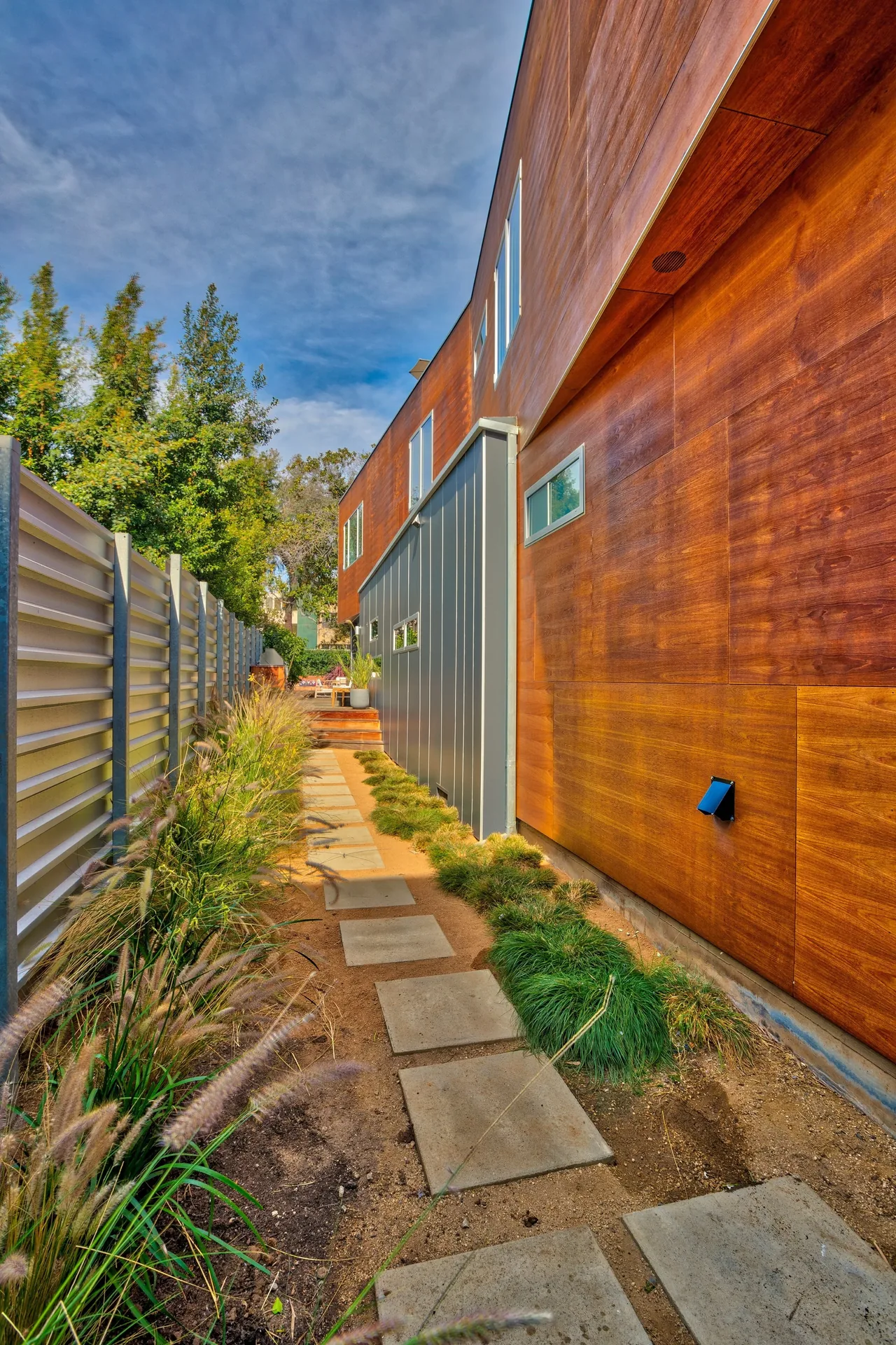 Side exterior pathway of a modern house with wood and metal panel siding and a landscaped garden bed.