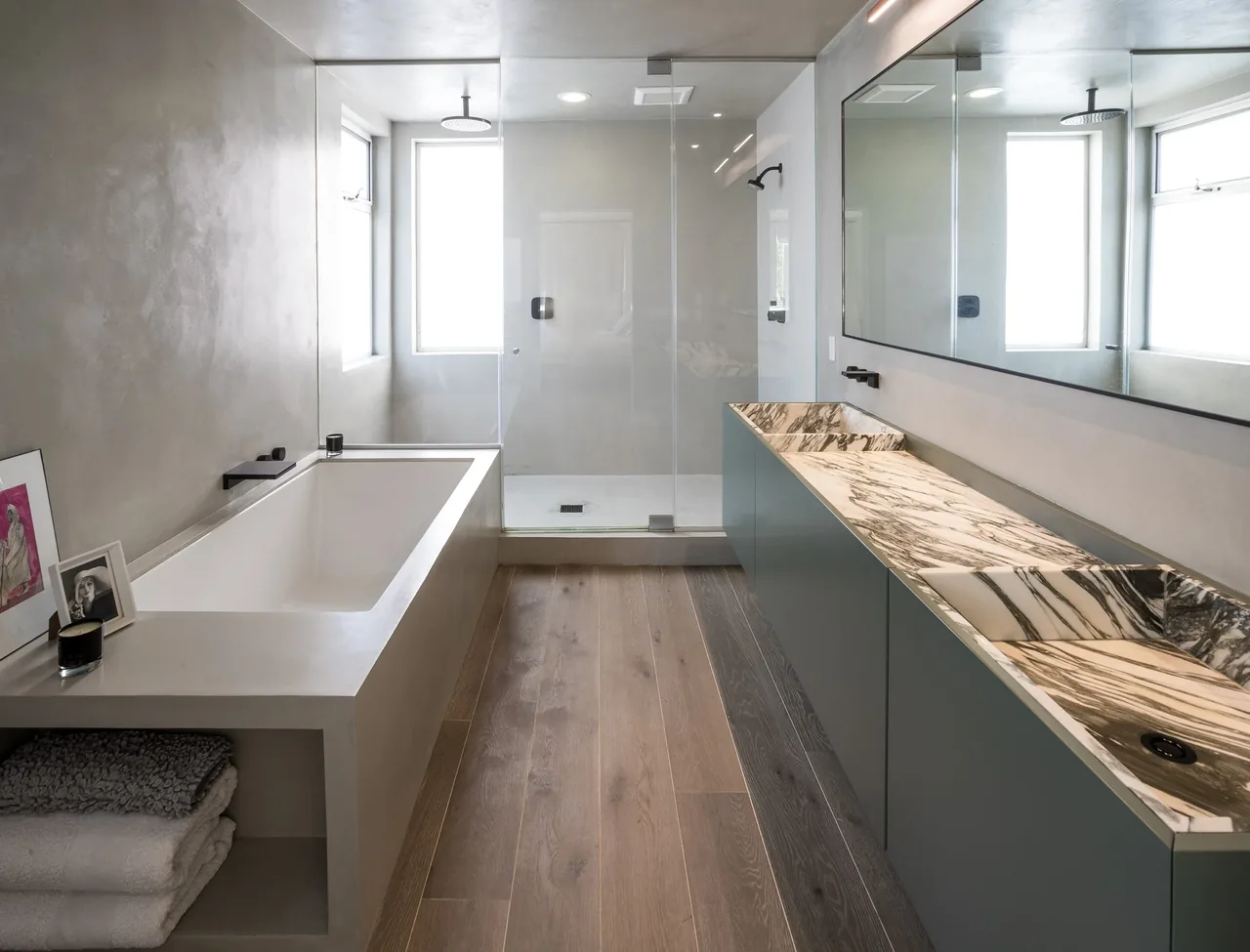 Bathroom features a built-in tub, dual marble sinks, wood flooring, and a frameless glass shower enclosure.