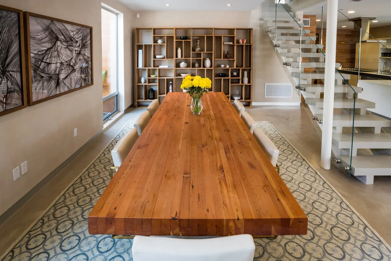 Dining room features a large wooden table, white upholstered chairs, an open shelving unit, and a staircase with glass railing.