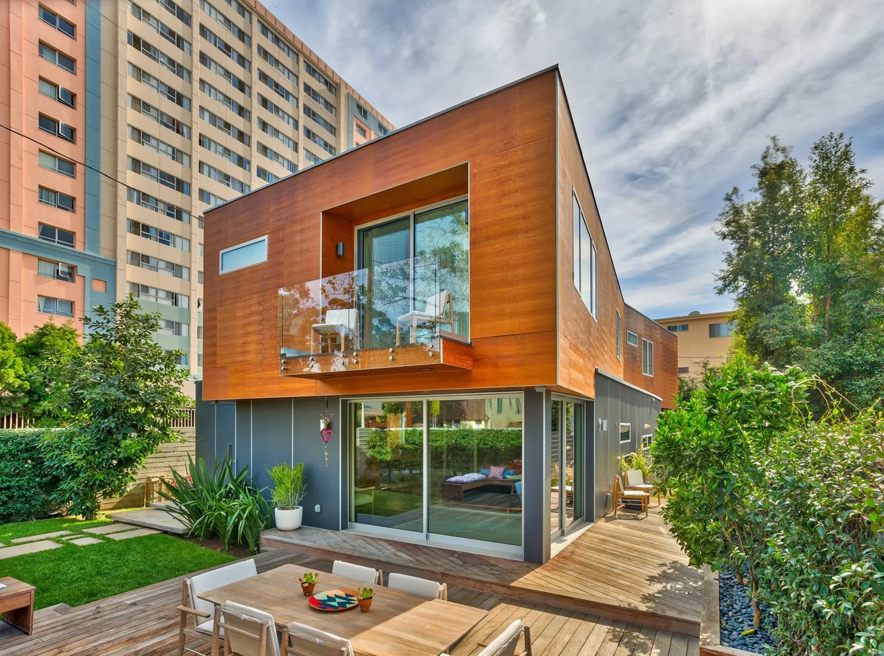 Two-story modern house with wood cladding on the upper floor, a glass balcony, and an outdoor deck with a dining table.