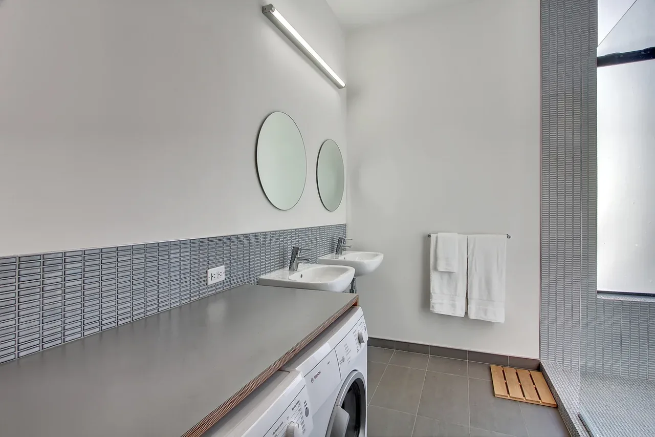 Laundry room features dual sinks with round mirrors, gray tile backsplash, and front-loading washer with gray countertop above.