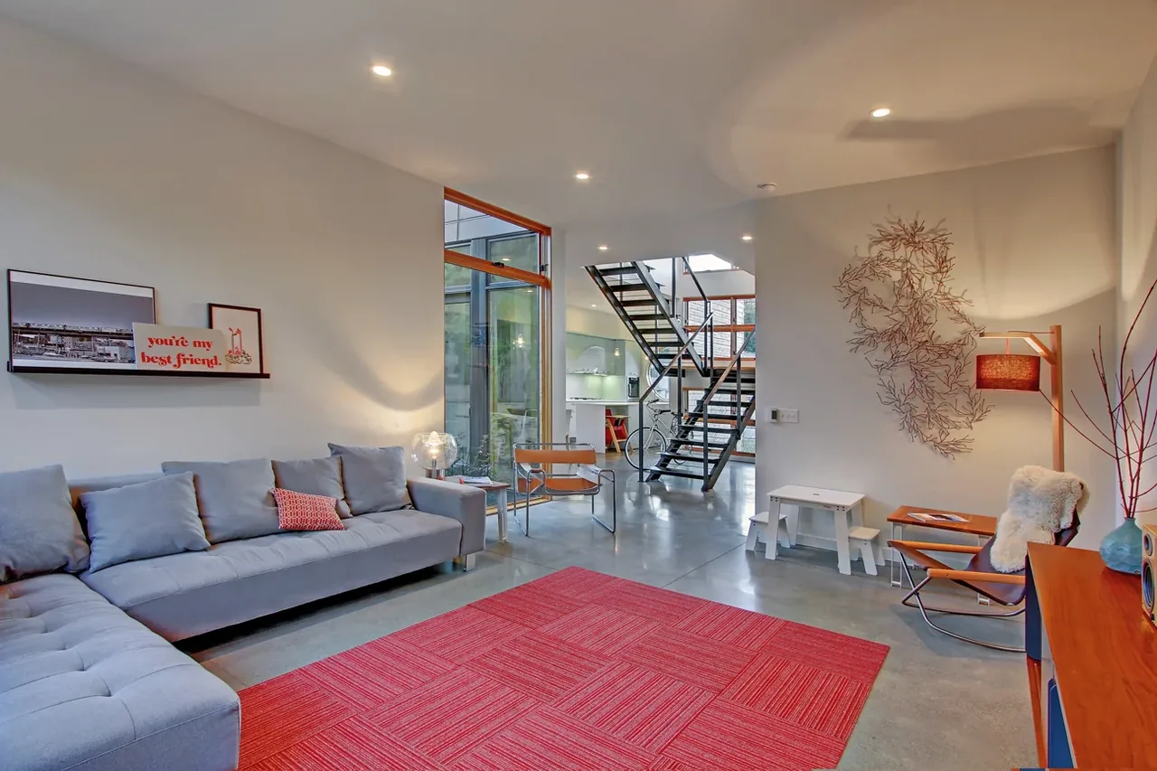 The living room features a gray sectional sofa and concrete floors with a metal railing staircase leading to an upper level.