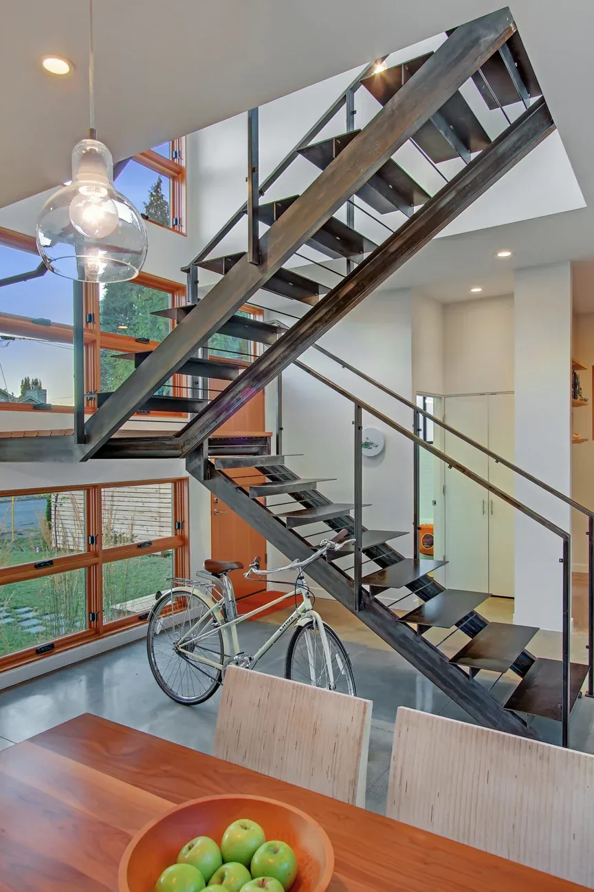 Modern black metal floating staircase next to wood-framed rectangular grid windows overlooking an entry area with a bicycle.