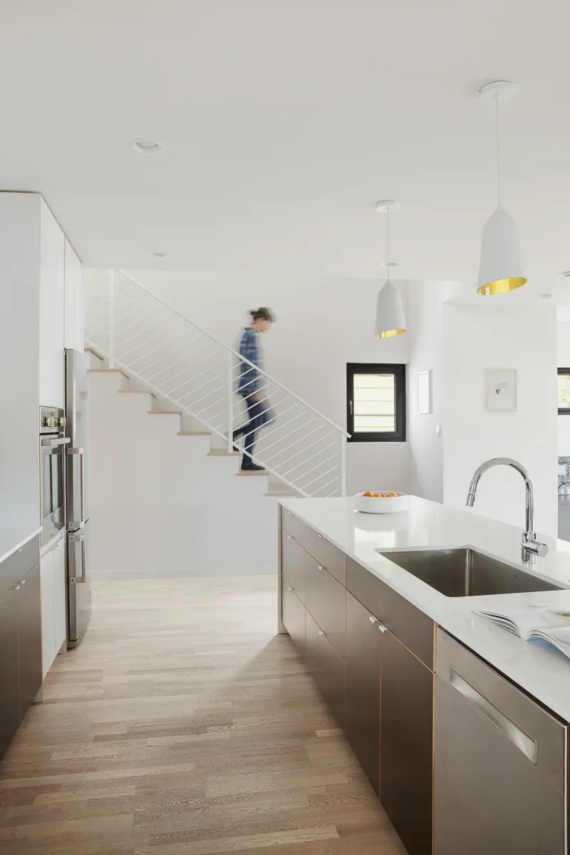 Minimalist kitchen with white countertops, wood cabinetry, and white pendant lights over the island, featuring light wood floors and visible stair railing.