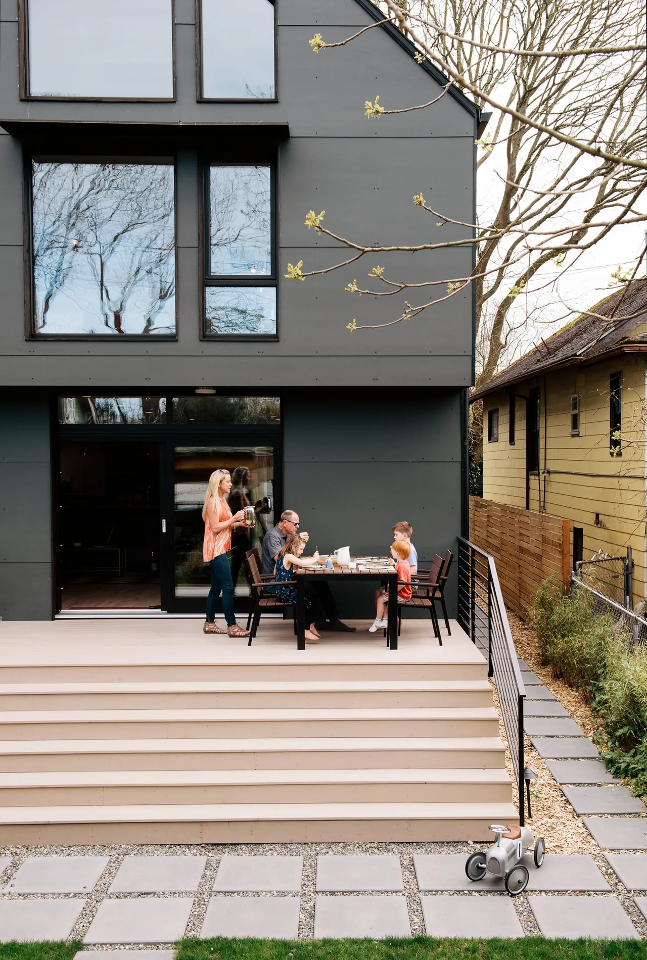 Back exterior of a modern dark gray house with a family dining on the wooden deck accessed by wide light-colored steps.