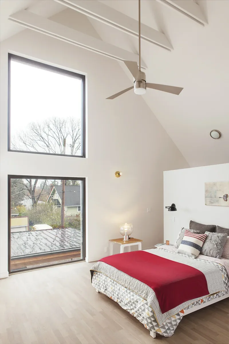 Bedroom with vaulted ceiling and large black-framed square windows overlooking exterior surroundings.