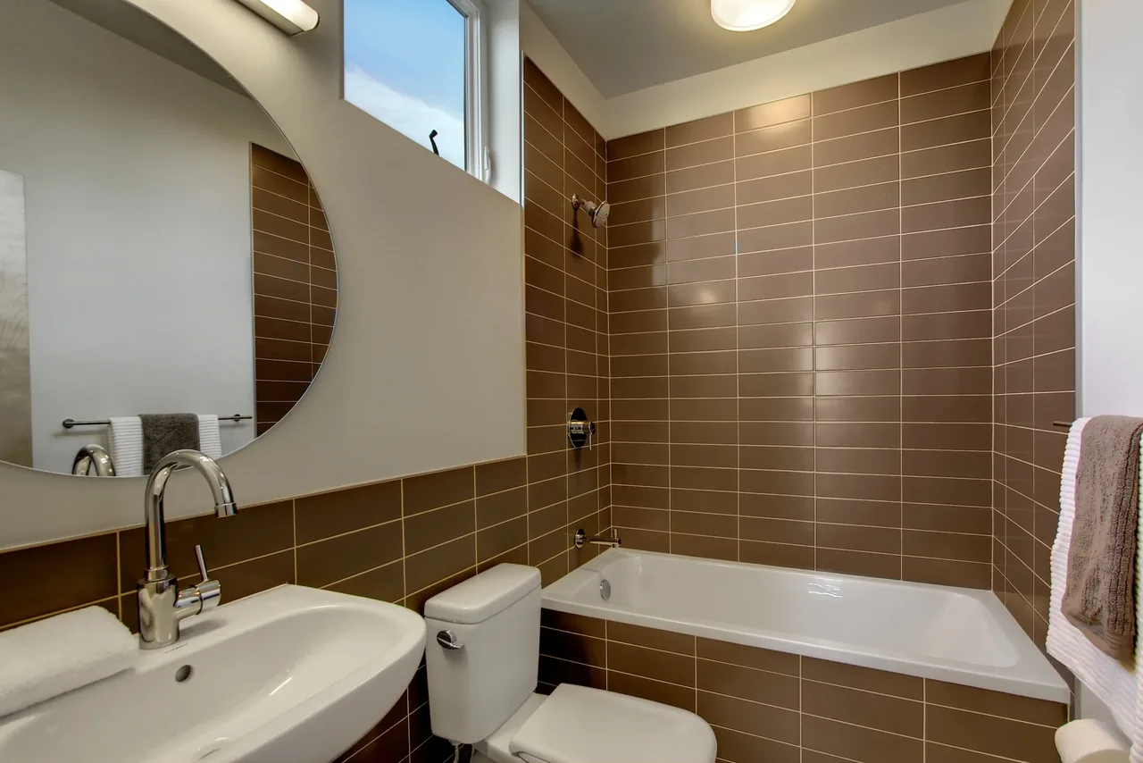 Bathroom features brown tile walls around the bathtub and a white sink with a round mirror.