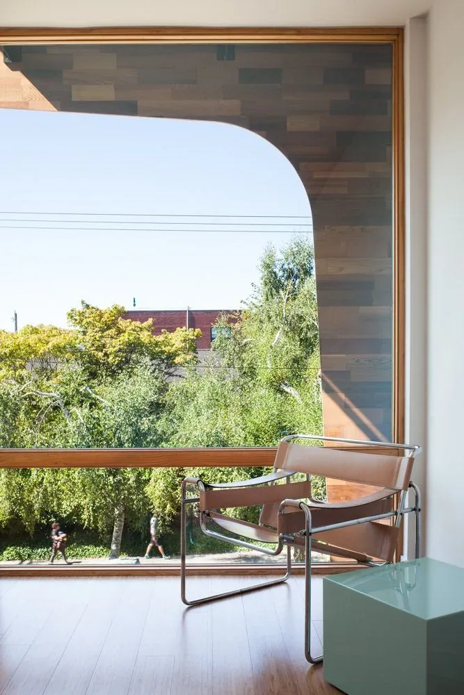 Interior view of a seating area with a leather and metal chair near a large wood-framed window overlooking trees.