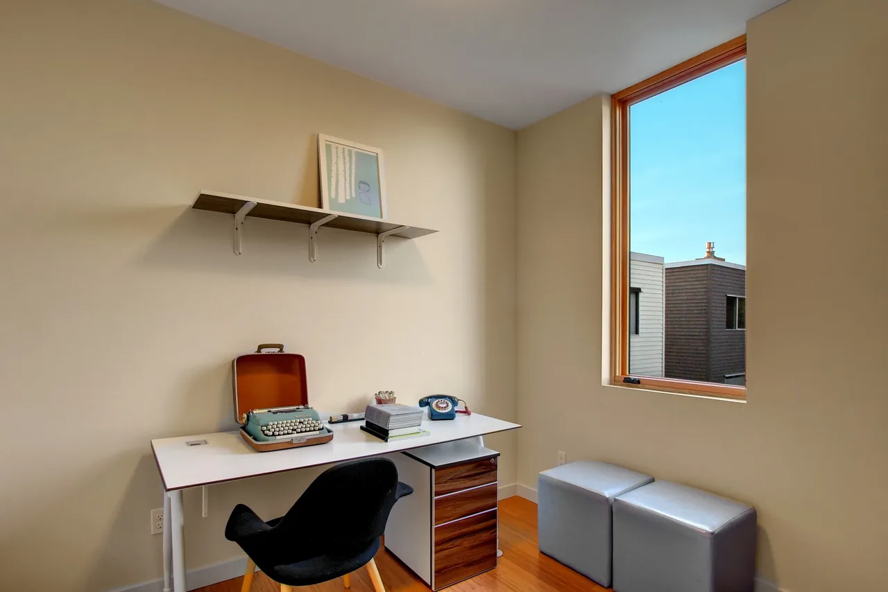 A minimalist office corner features a white desk with a typewriter and a vintage phone under a large window.