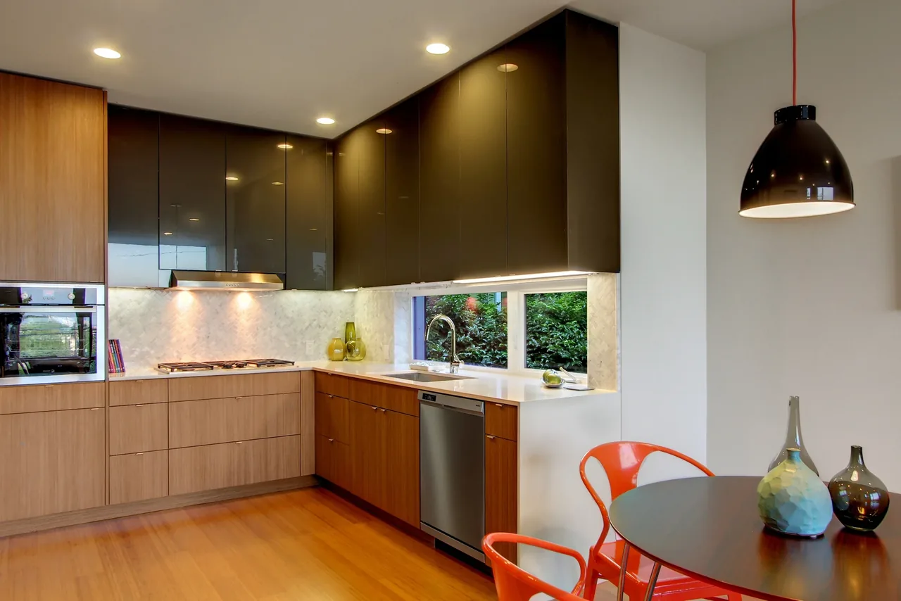 Kitchen features wood lower cabinets, dark upper cabinets, a white countertop, and a window above the sink with greenery outside.